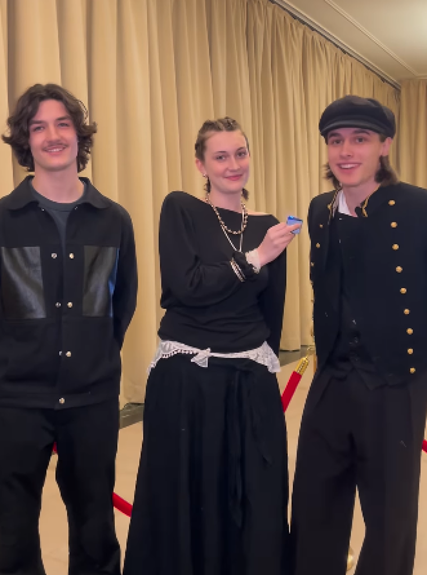 Three young adults stand side-by-side against a tan curtain backdrop, the center person holding a small blue object.