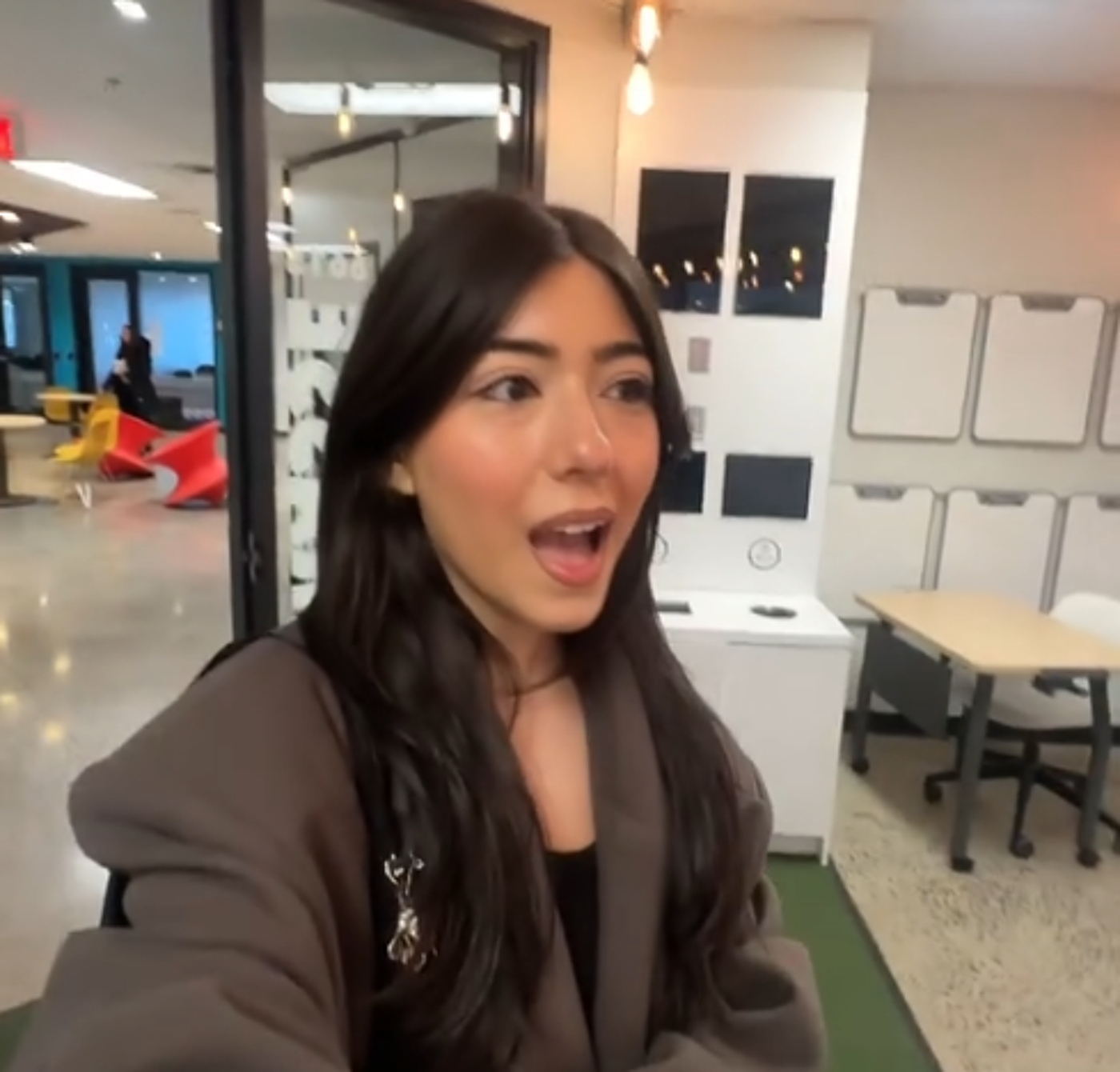 A young woman with long dark hair, wearing a brown coat, stands in an office space, speaking animatedly.