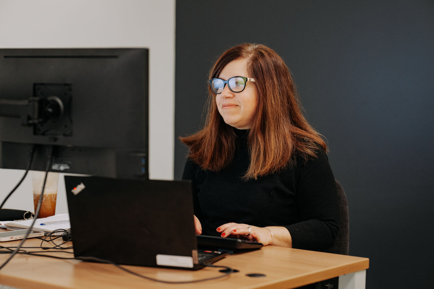The image features a woman sitting at a desk, using a laptop computer and wearing glasses.
