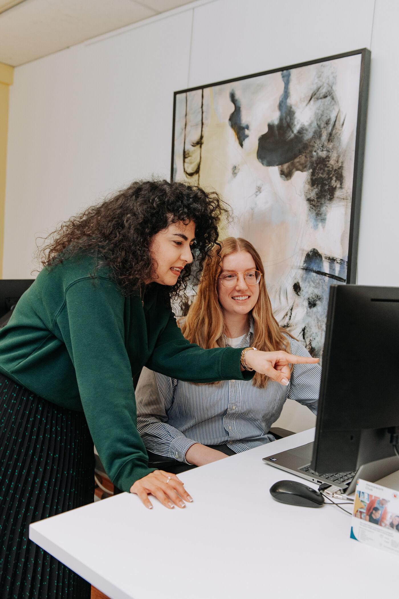 In the image, there are two women standing in front of a computer, with one pointing at the screen. They appear to be discussing or showing something on the computer.