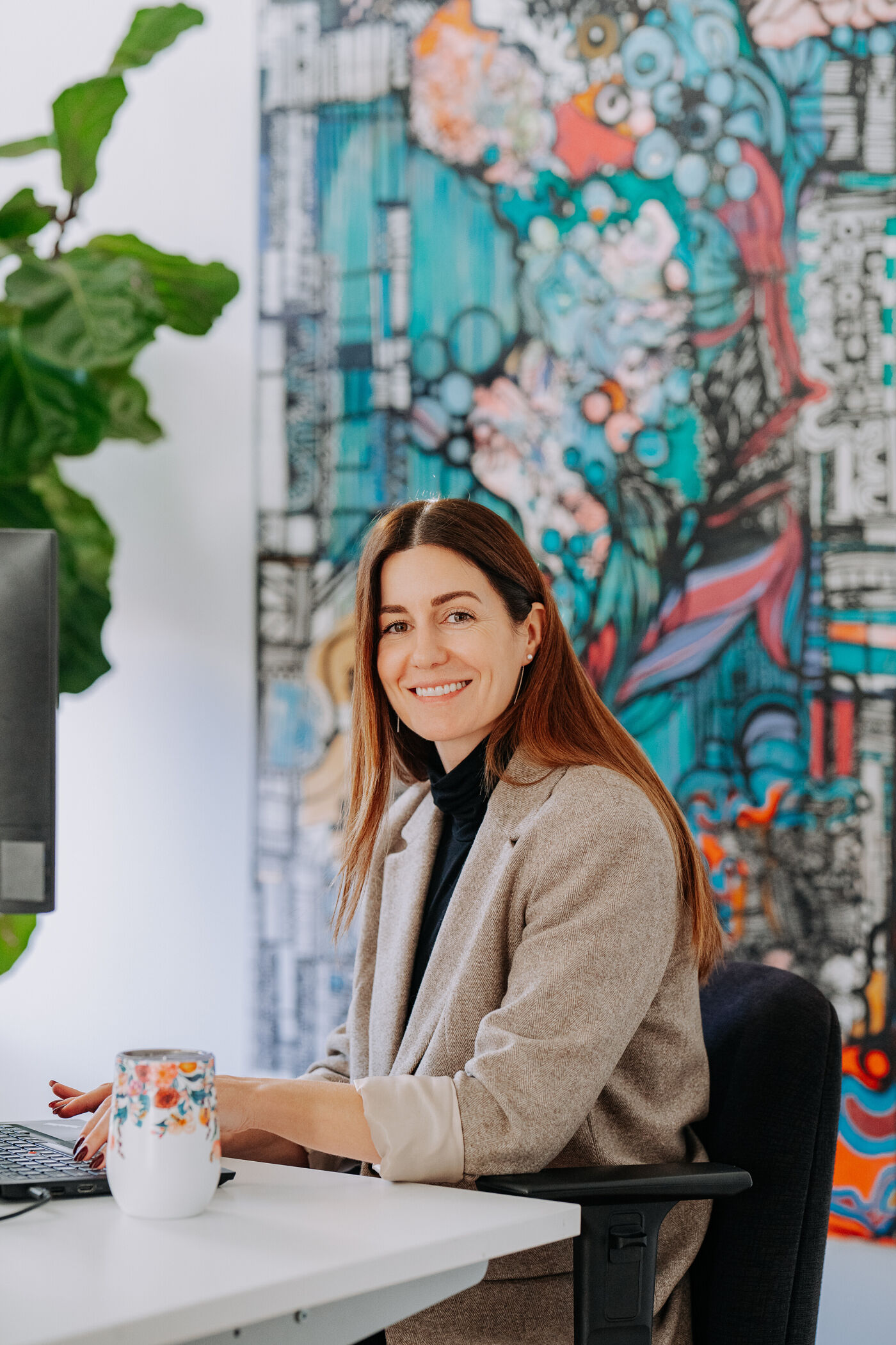In the image, a woman is sitting at a desk in front of a computer, smiling and holding a cup.