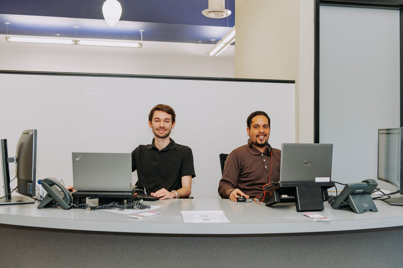 In the image, there are two men sitting at a desk with laptops, working together in a room.