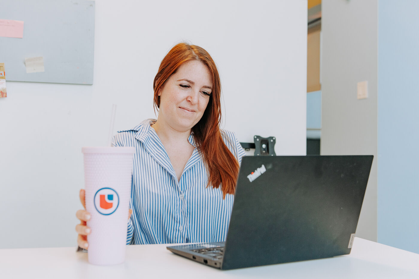 In the image, a woman is sitting at a table, smiling while using a laptop and holding a pink drink.
