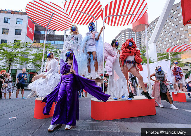 Models in avant-garde outfits pose on an outdoor stage with bold striped backdrops, creating a dynamic fashion display.