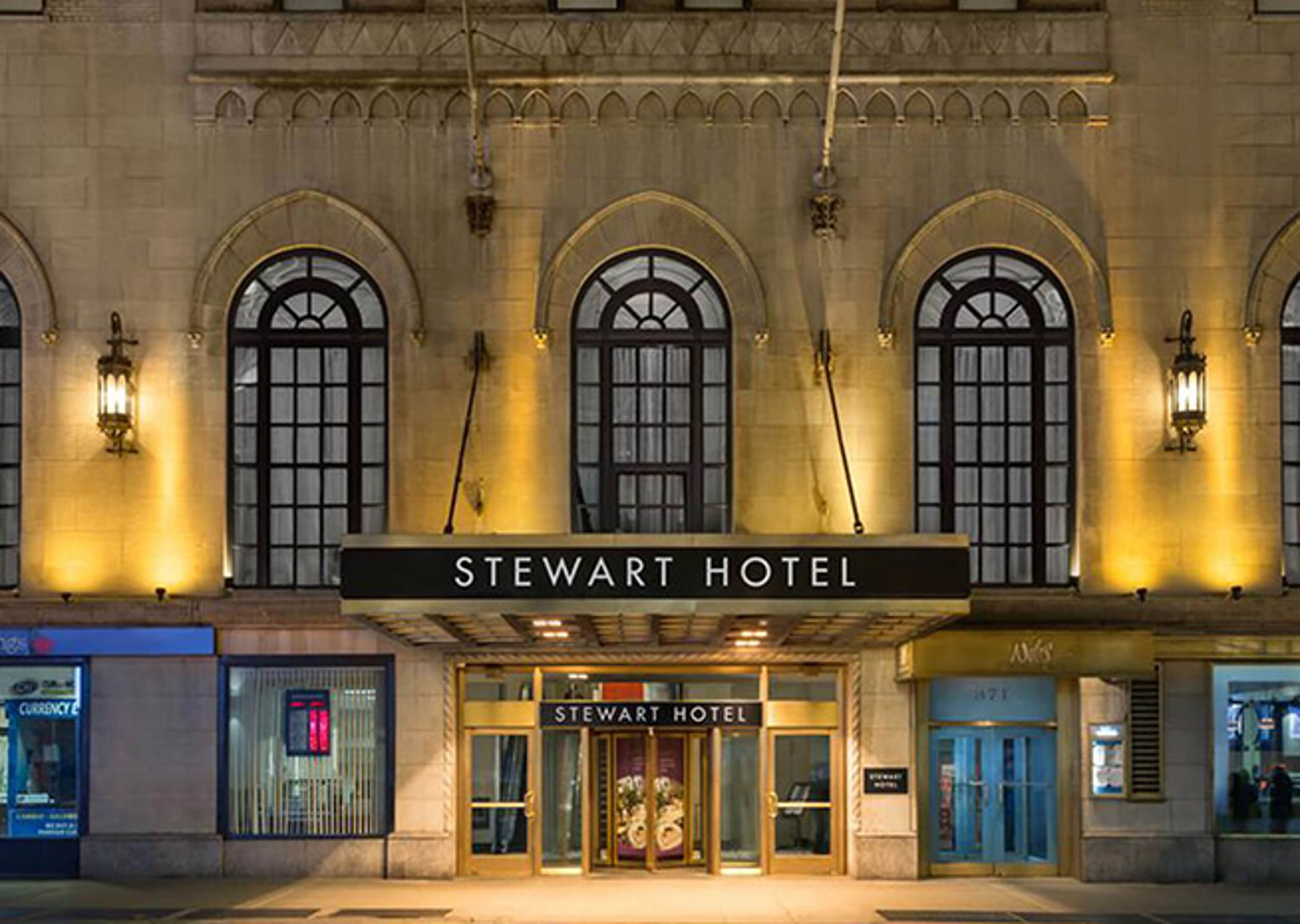 L'entrée illuminée de l'Hôtel Stewart, mettant en valeur son architecture classique avec des fenêtres voûtées et une esthétique vintage au crépuscule.