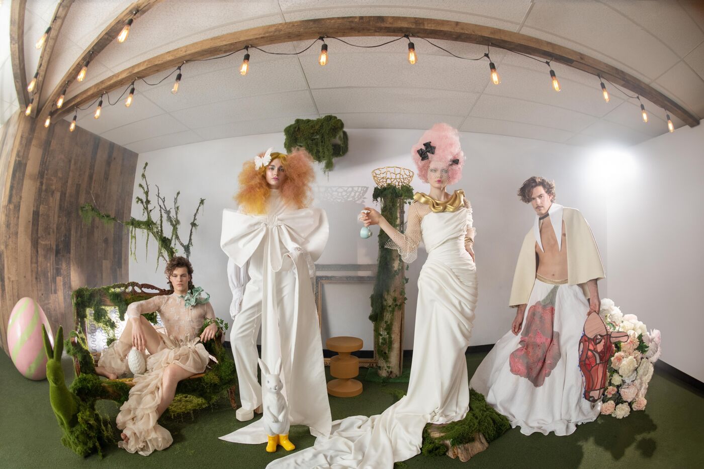 Four models in whimsical Easter-themed attire pose in a studio setting adorned with greenery and pastel colors.