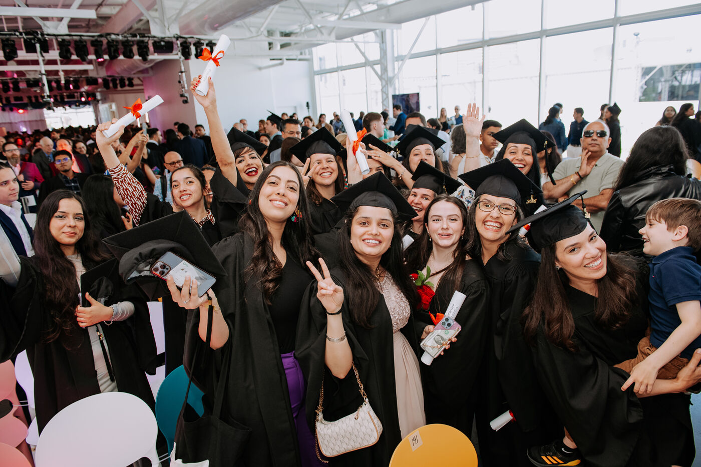 A large group of graduating students in black graduation gowns and caps are densely packed together, many holding scrolls.
The students are diverse in ethnicity and have varying hairstyles and levels of makeup. Some are smiling directly at the camera, while others are looking at each other or off to the side.
A few students in the foreground are holding small bags or purses. Behind the graduates, other people, possibly family and friends, are visible but less distinct. The background shows a bright, modern interior with large windows letting in natural light. A few people in casual clothes are seen in the distance.
A young boy in a blue shirt is being held up near the right edge of the image, partially obscured by one of the female graduates.
The overall mood is joyful and celebratory, typical of a graduation ceremony.