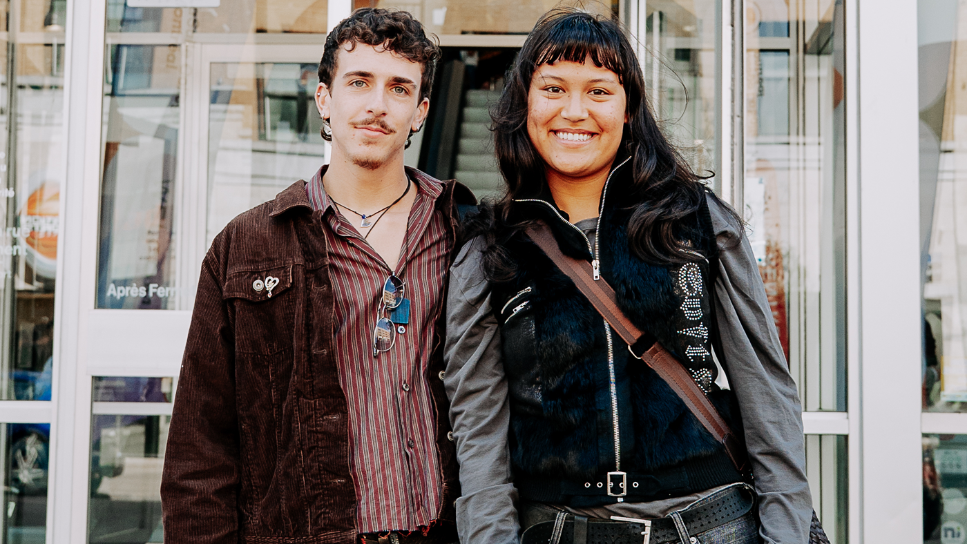 Two young people are smiling at the camera in front of a building entrance. The person on the left is wearing a brown jacket and a striped shirt, while the person on the right is wearing a dark vest and a grey shirt.