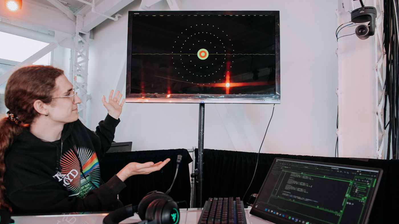 A woman with curly hair is presenting a screen displaying a visual representation of sound waves. The setup includes a keyboard, headphones, and a laptop with code visible on the screen, suggesting an audio engineering or music production environment.