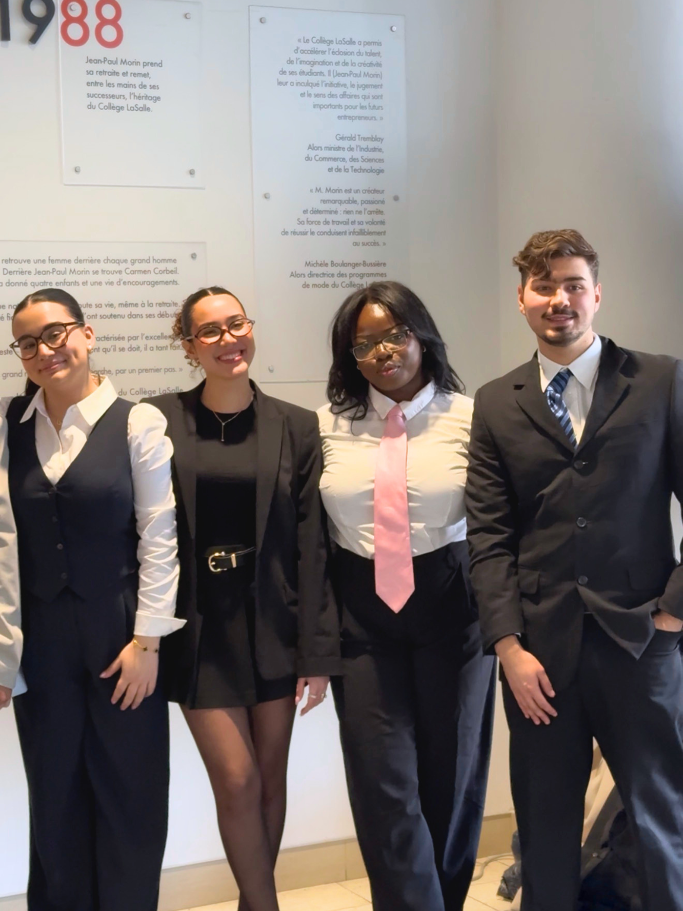 Four young students, three women and one man, smile at the camera, dressed in formal attire, with a commemorative plaque in the background.