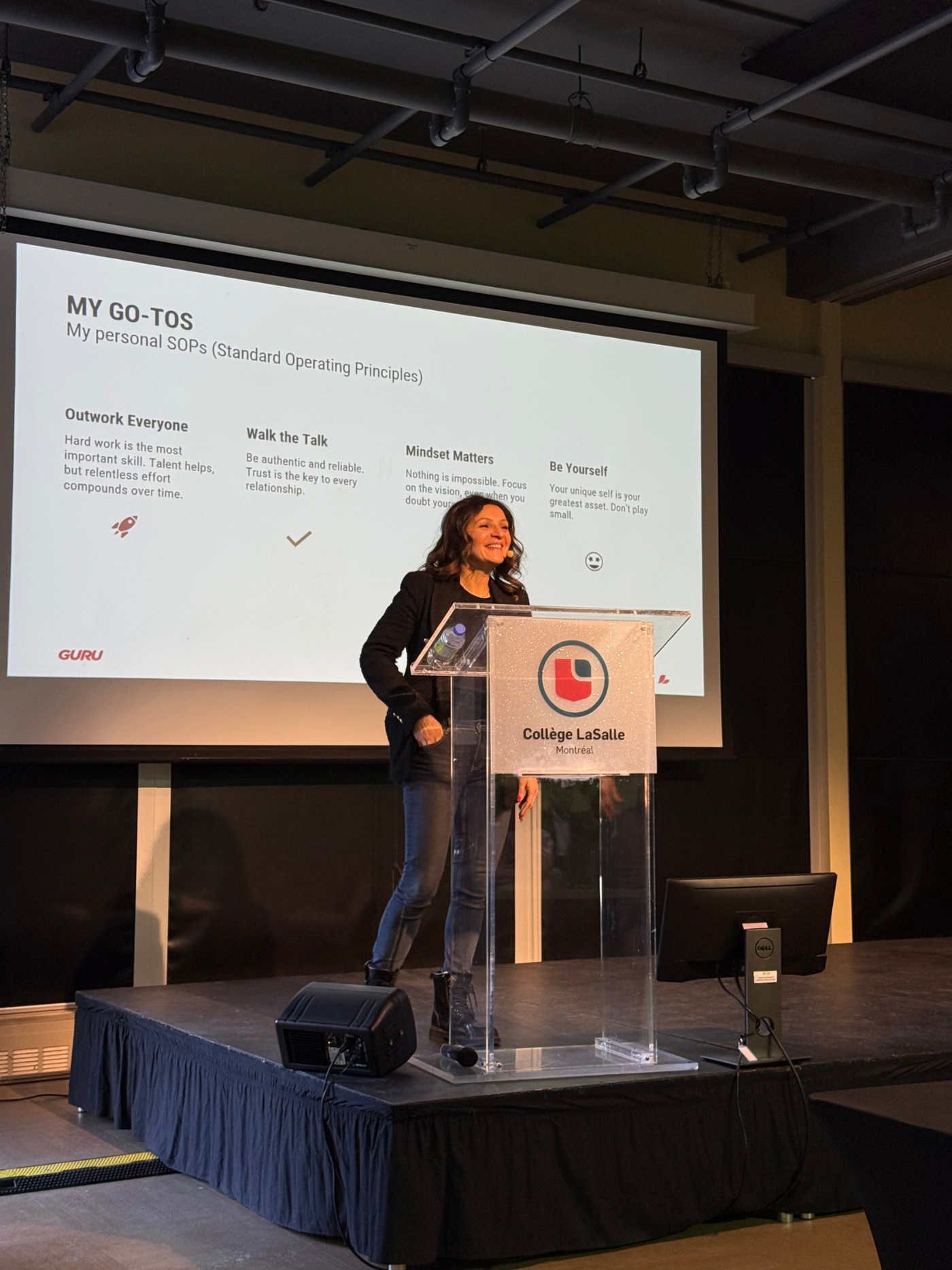 A woman smiles while speaking on a stage in front of a large screen displaying her personal standard operating principles, titled "MY GO-TO-S".