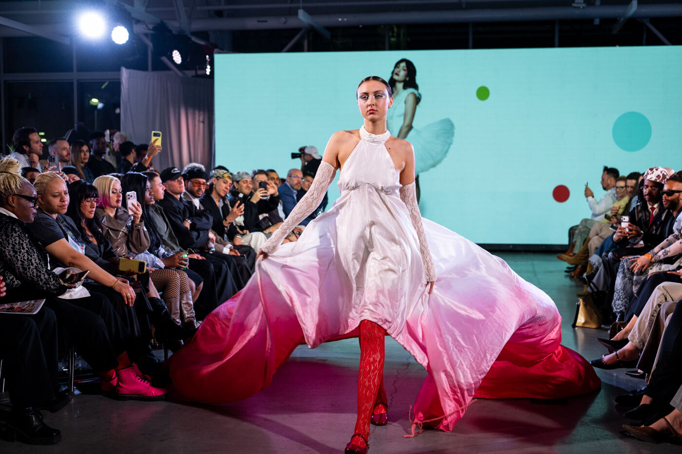 A model walks the runway in a white and pink gown with a long train, paired with red stockings and long gloves, in front of an attentive audience.
