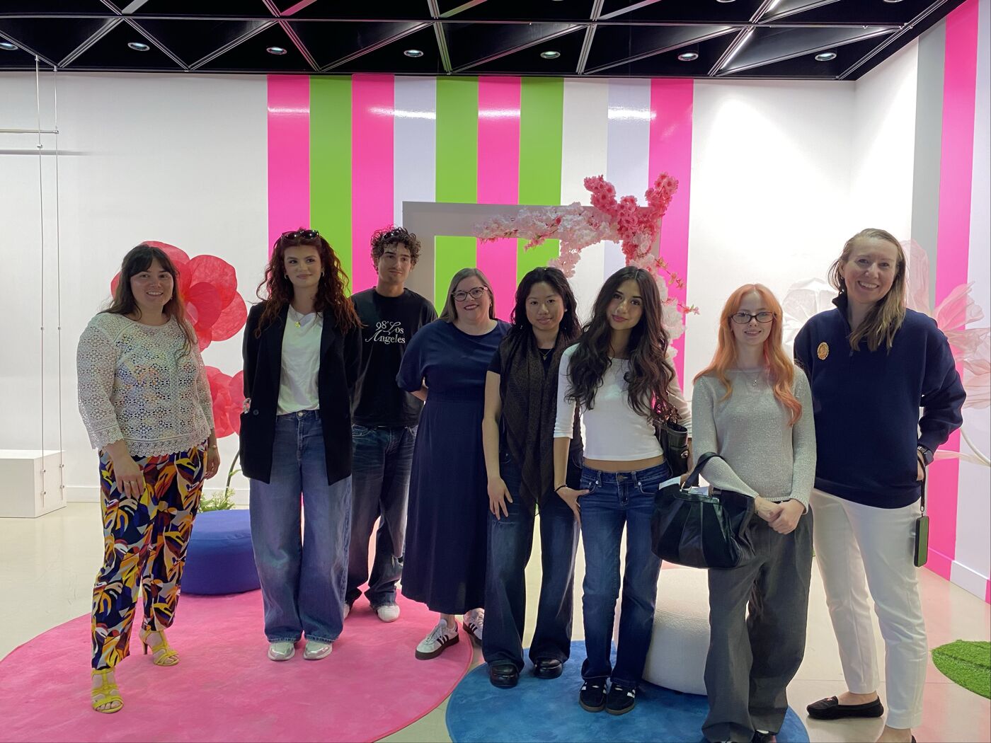 Group of eight people standing in a bright, modern interior with bold pink, green, and white wall panels, circular floor accents, and decorative floral elements.