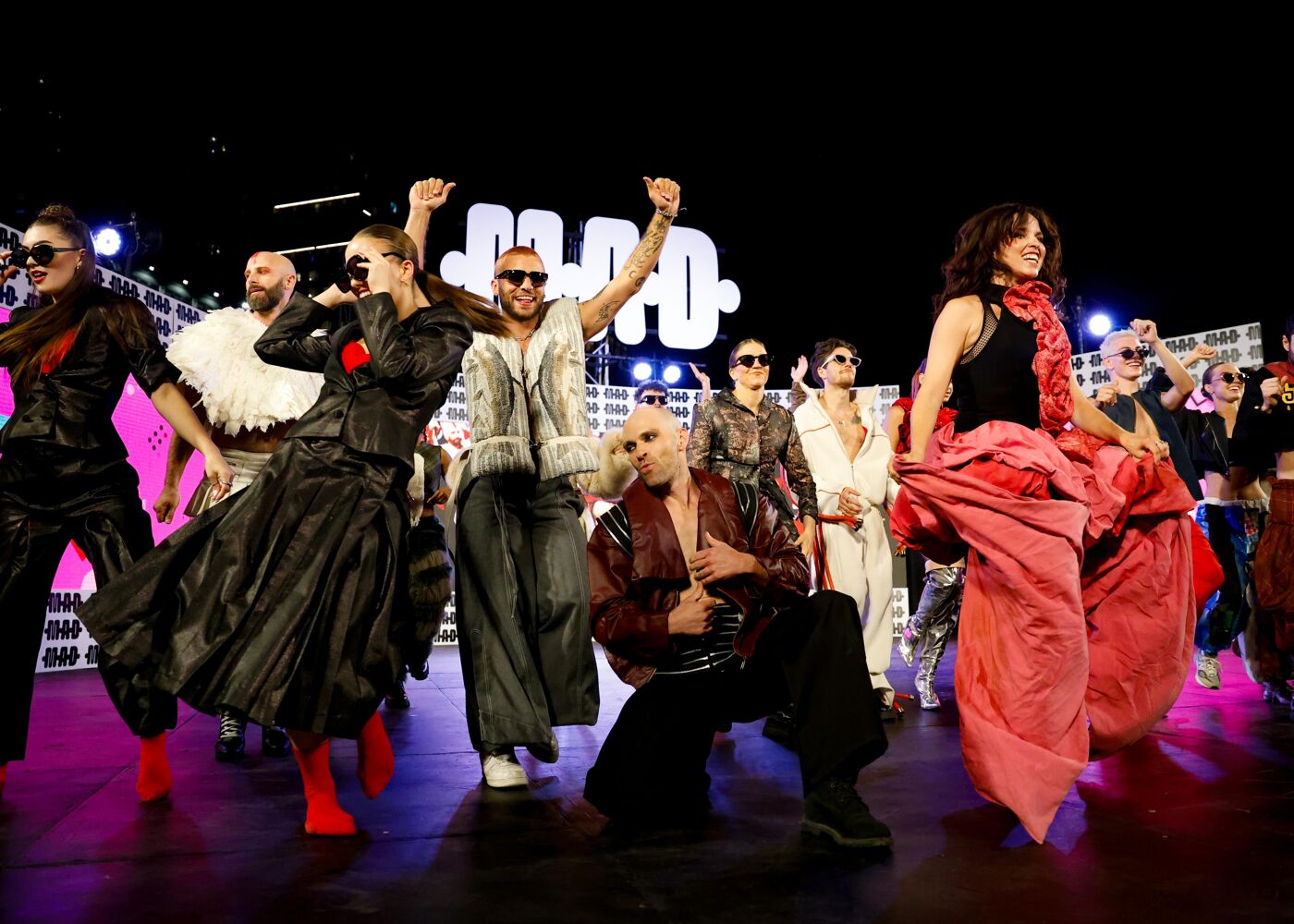 A group of people in flamboyant outfits and dramatic makeup pose on a brightly lit stage, possibly during a fashion show or performance.