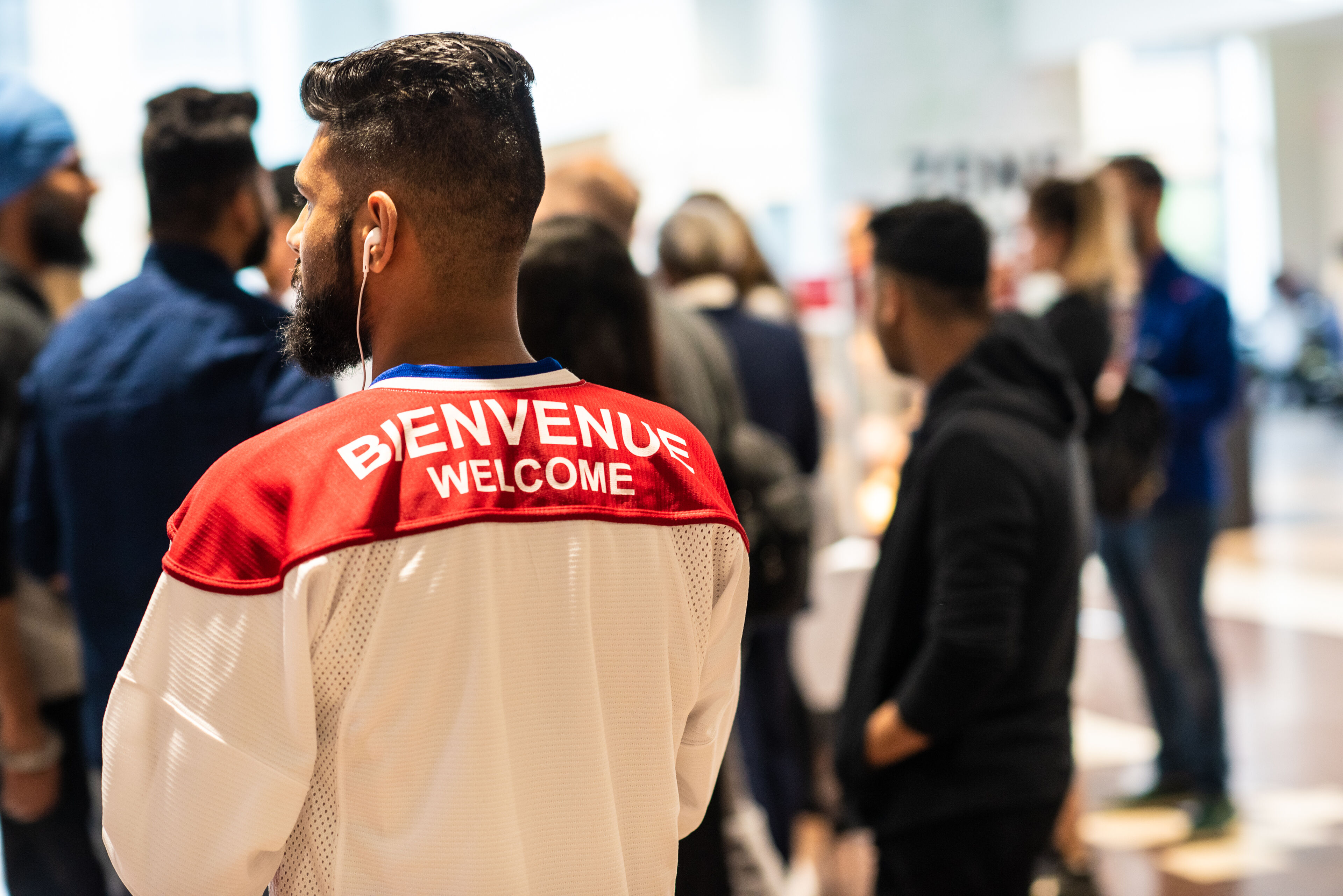 A man wearing a jersey with 'Bienvenue - Welcome' stands amidst a crowd, symbolizing a warm, multilingual greeting.