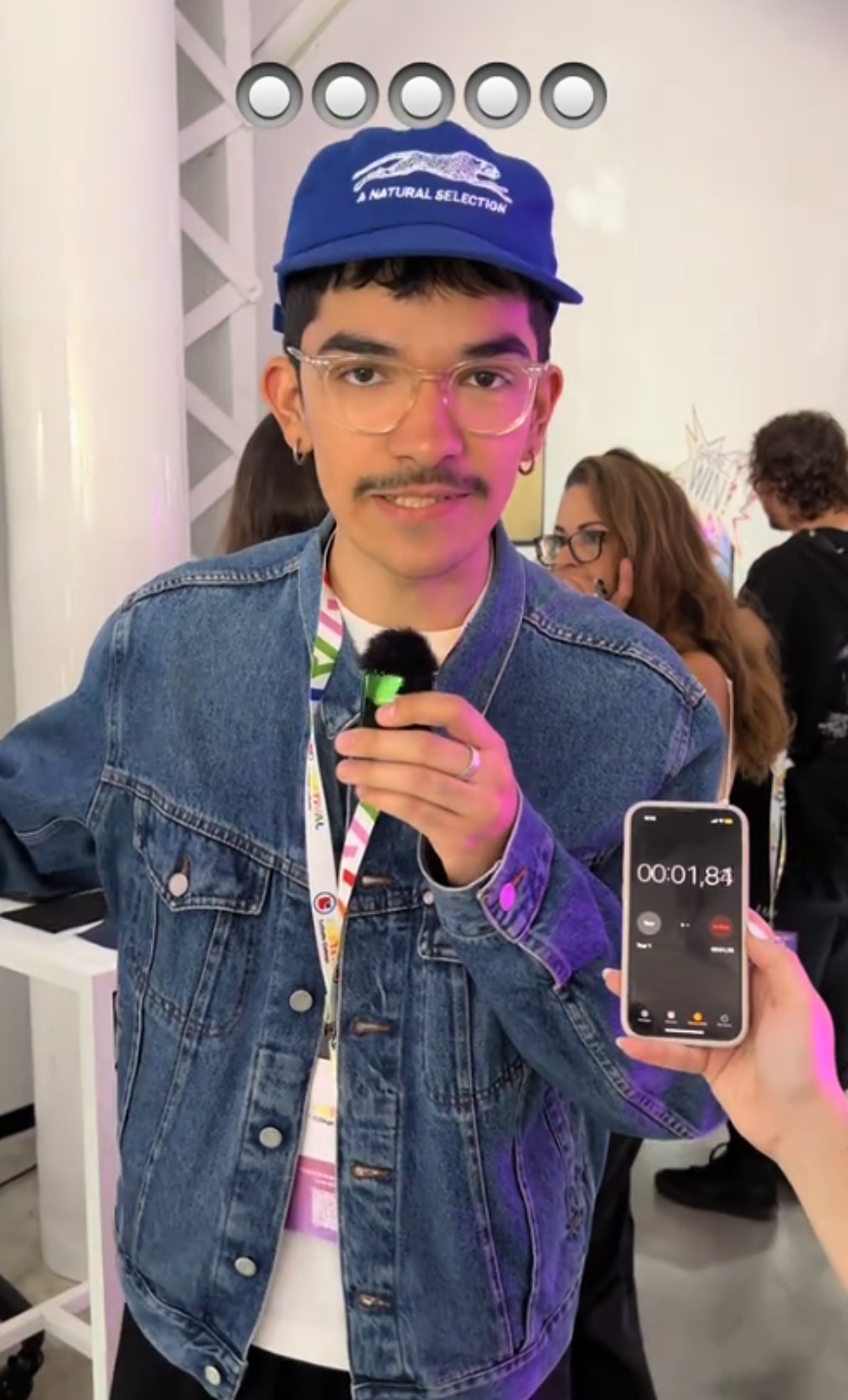 A young man with glasses and a mustache is holding a microphone and wearing a denim jacket and a blue cap. Someone is holding a phone displaying a timer. There are people in the background and white architectural elements.