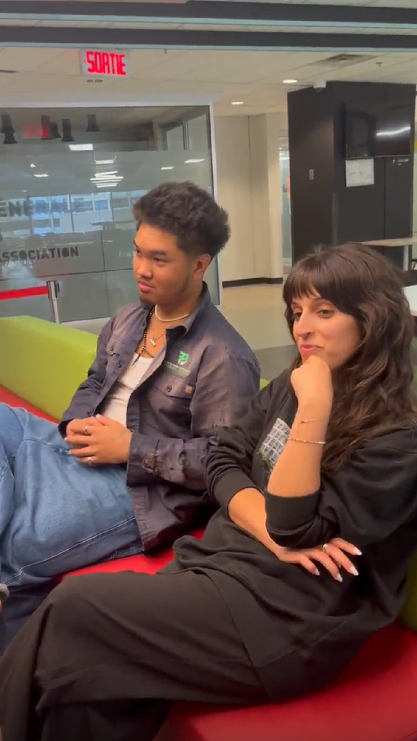 Two young adults sit on a colorful couch in a modern lounge, appearing relaxed and engaged in conversation. The setting features glass walls, indicating a public or educational space, with the word "SORTIE" visible, suggesting a French-speaking environment.