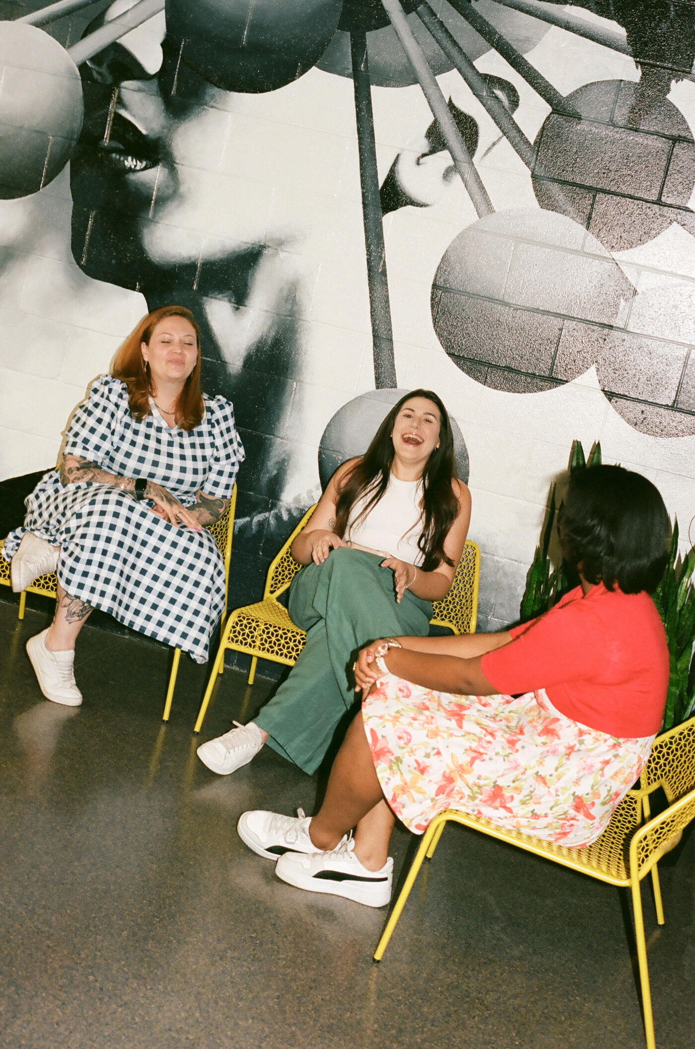 Three women enjoying a lively conversation, seated on yellow wire chairs against a black and white mural backdrop.