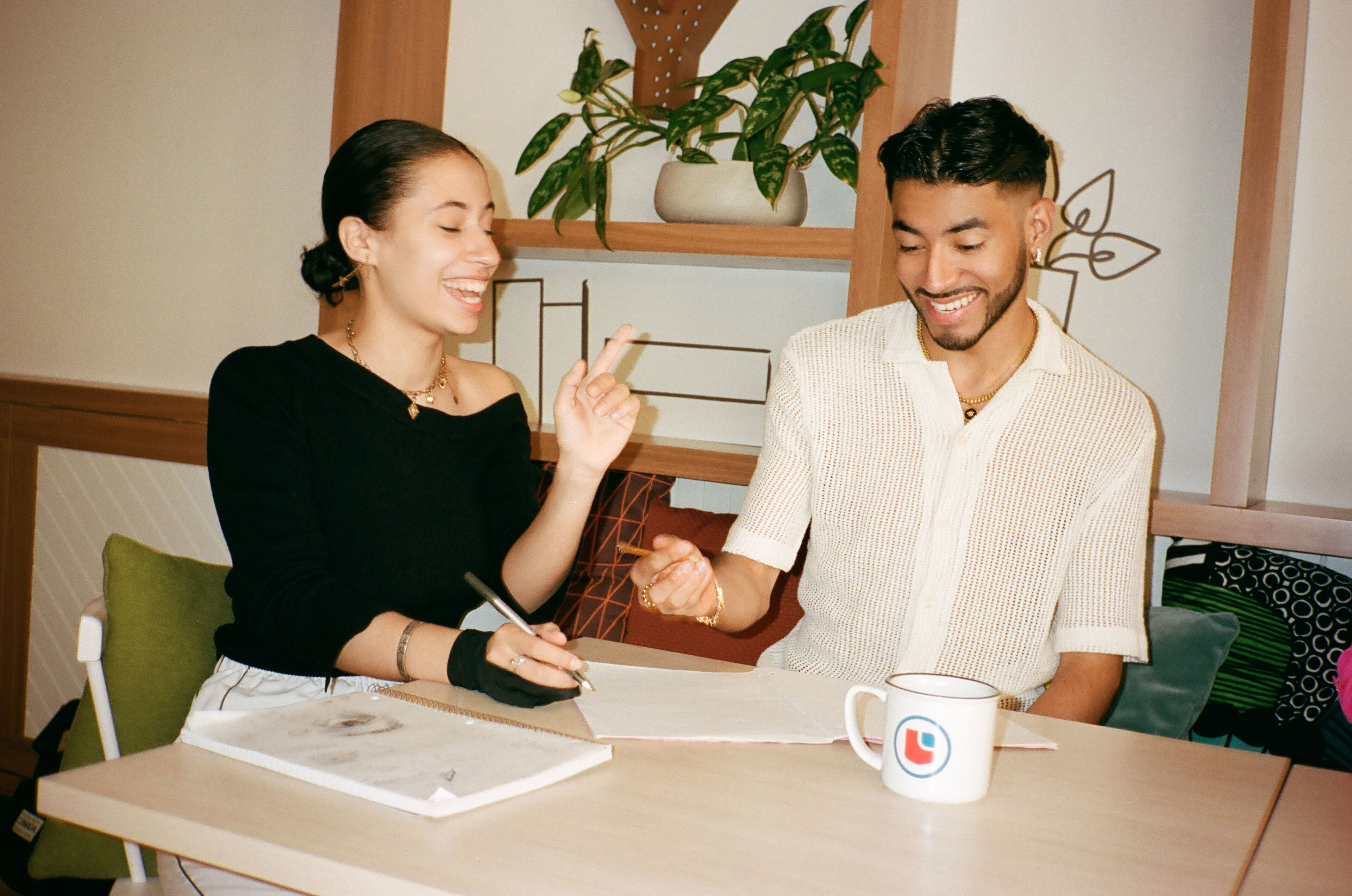 Two smiling friends enjoy a moment of creativity, sketching together in a sunlit room.