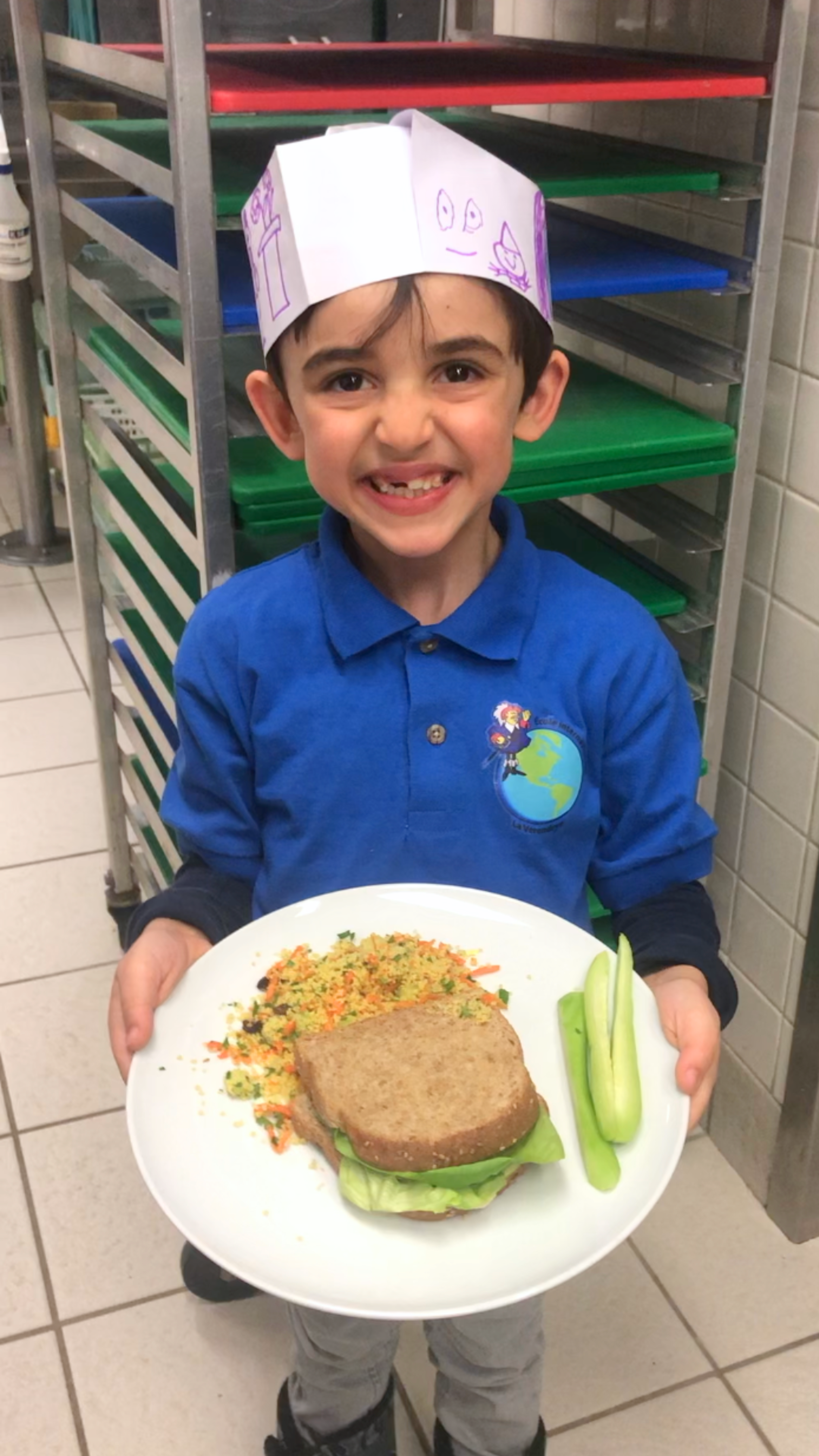 A smiling young boy in a chef's hat presenting a homemade sandwich with veggies on a plate, exuding culinary excitement.