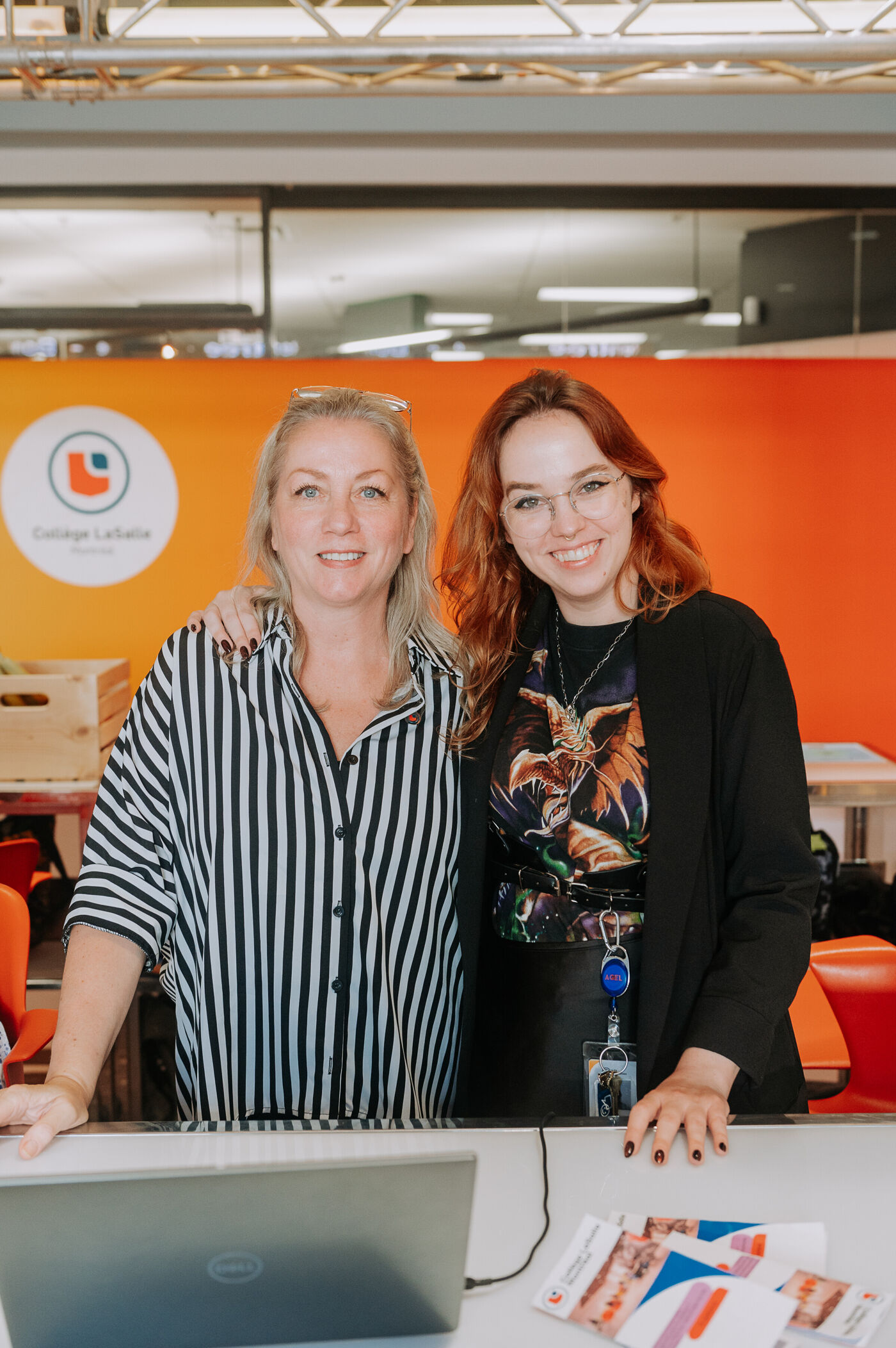 Two smiling women stand together at Collège LaSalle, with an orange background and a logo visible. They appear professional and approachable, wearing casual business attire. A laptop and brochures on the table suggest an educational or work-related setting.