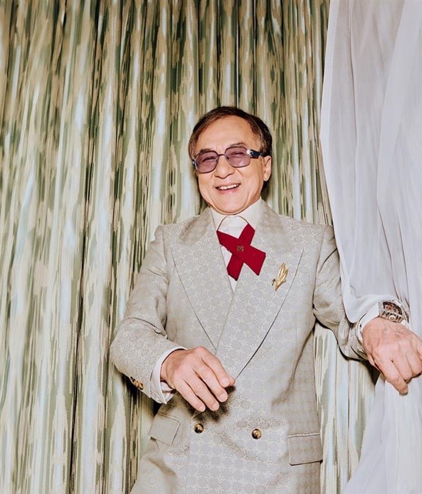 The image shows a smiling man in a light-colored suit with a red cross-shaped accessory on his chest. He is wearing glasses and a watch, and he is standing in front of a patterned curtain.