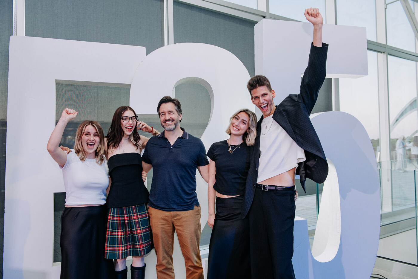 A group of five people are posing in front of large white letters, seemingly celebrating an anniversary or milestone. They are smiling and appear to be in a cheerful mood, with one person raising their fist in the air.