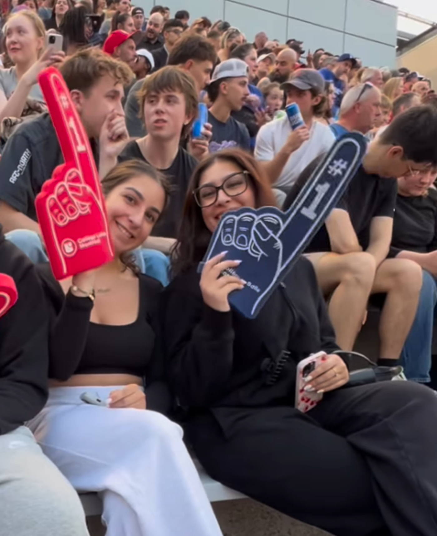Two young women, one wearing white pants and a black top, and the other in black pants and a black top, are seated in a crowd of people at an outdoor event, each holding up a large foam number one hand.