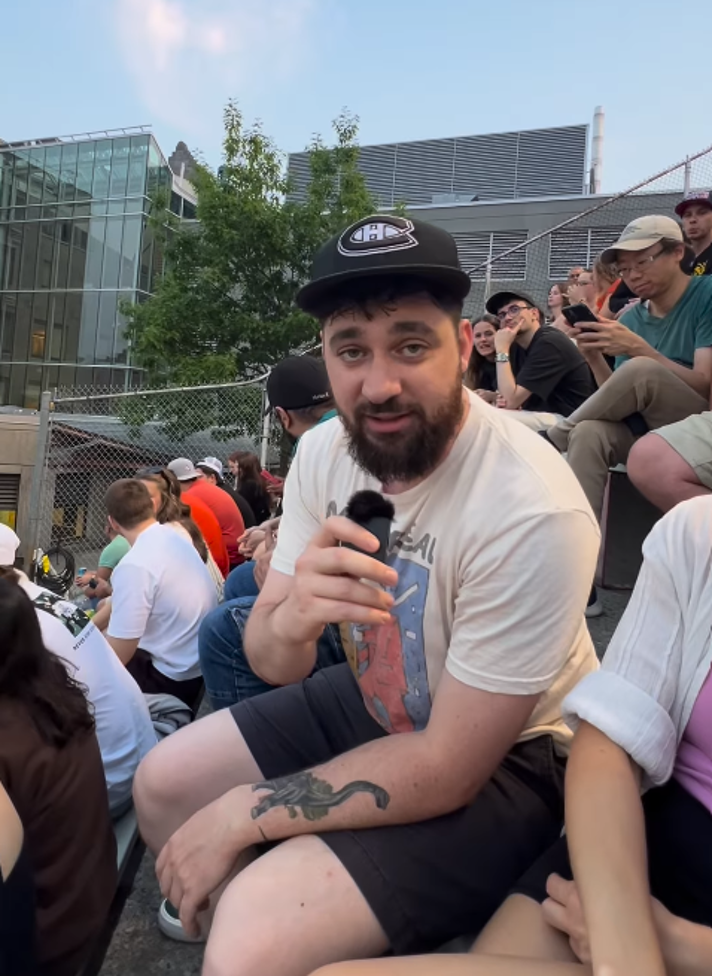 A man with a beard and a baseball cap sits outdoors holding a microphone, surrounded by a crowd of people.