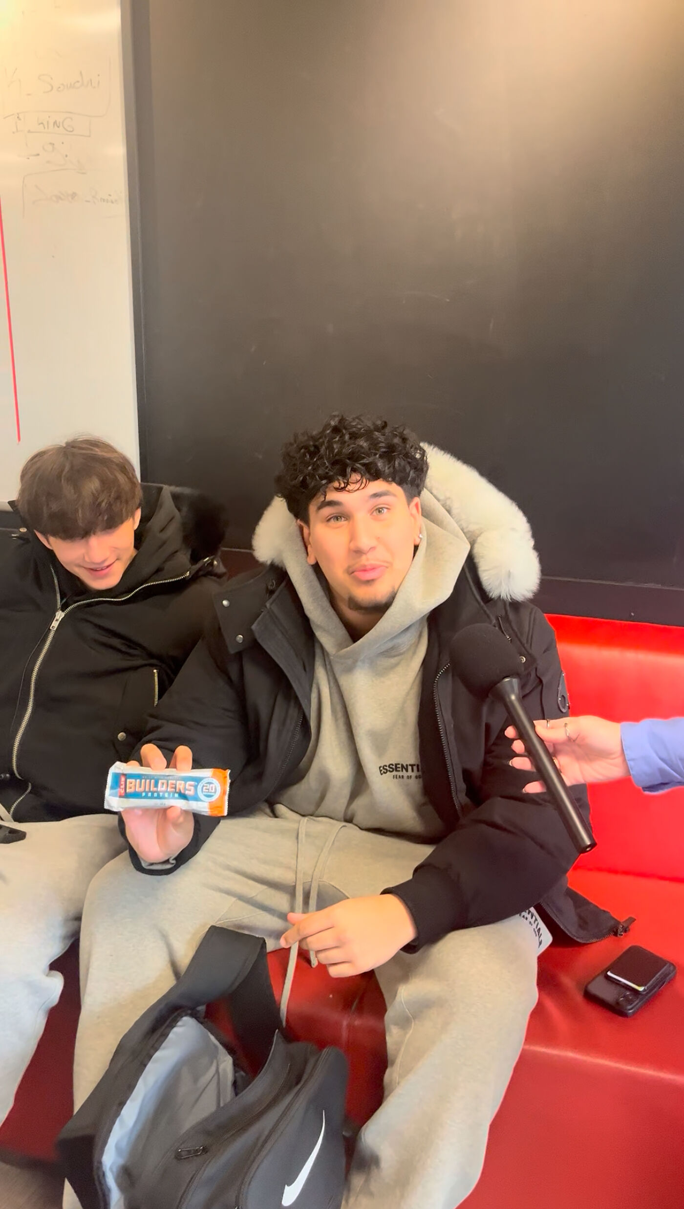 In the image, a man is sitting on a red couch, holding a candy bar, and being interviewed by a reporter.