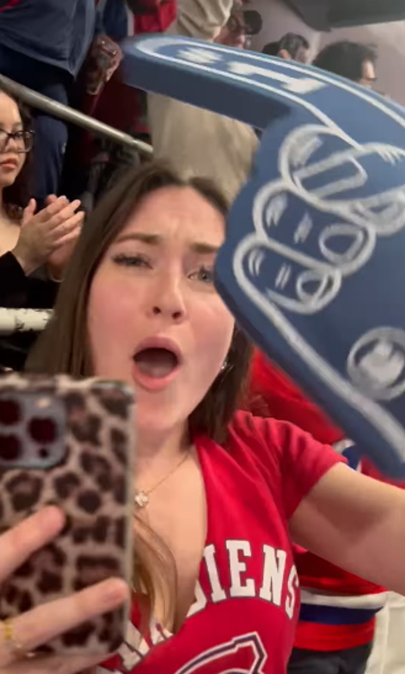Close-up view of a young woman with long brown hair, wearing a red jersey with white lettering, holding a phone with a leopard-print case while a large foam hand obscures part of her view.