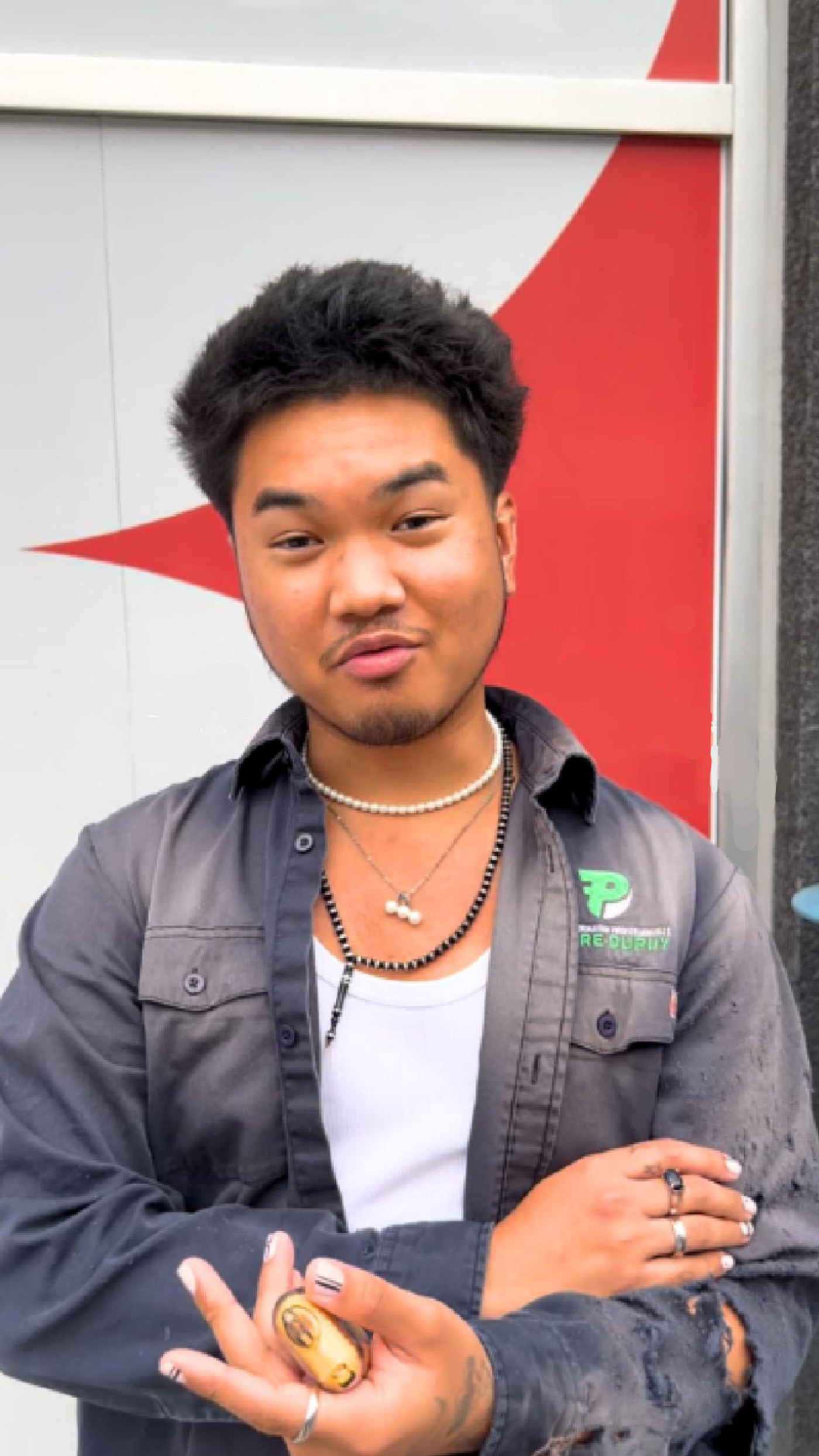 A young man poses confidently in a casual outfit, wearing layered necklaces, rings, and a dark shirt over a white tank top. He holds a small item in his hand and has painted nails, expressing a unique, edgy style. The red and white background adds a modern touch.