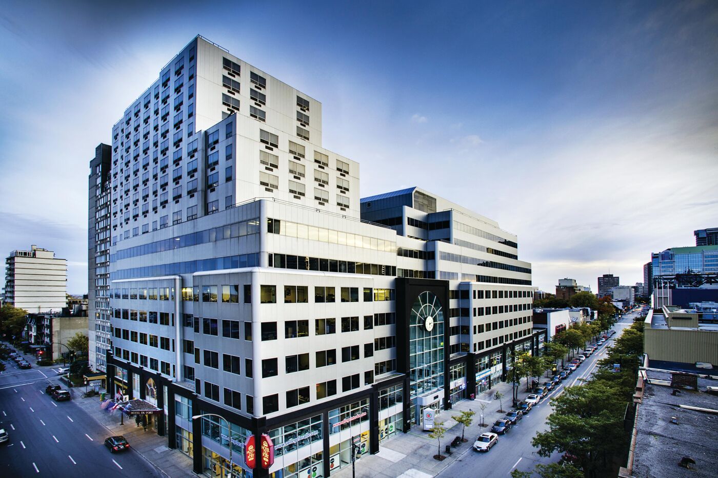 A modern office building with a glass and metal facade, situated at an urban intersection with traffic and trees lining the sidewalk.
