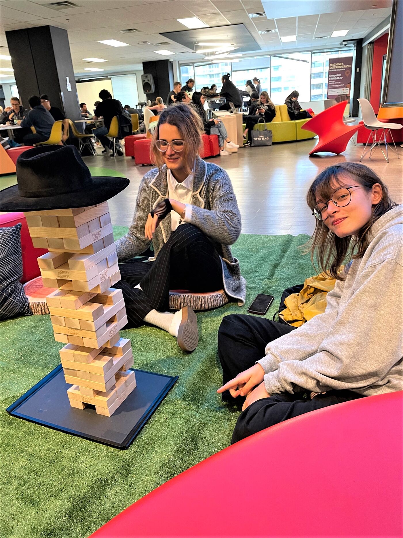 Two young women sit on a green artificial turf floor in a modern office space, playing Jenga with a large wooden tower, surrounded by colorful beanbag chairs and other furniture, while other people are visible in the background.