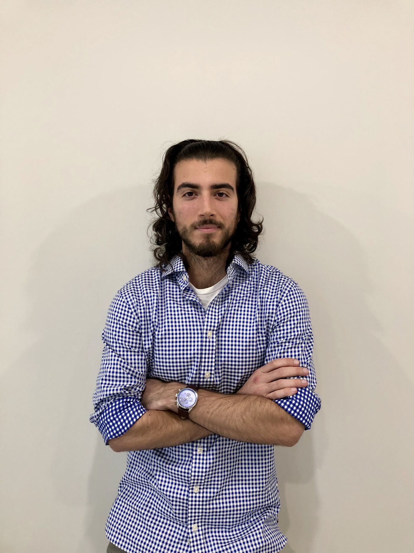A man with long hair and a beard poses confidently against a plain background, wearing a blue and white checked shirt and a wristwatch.