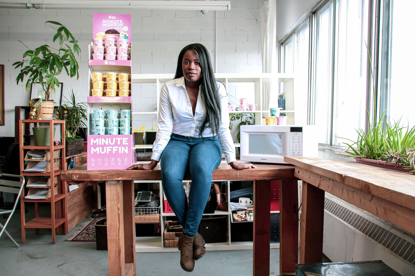 A woman entrepreneur sits confidently on a wooden table in a bright workspace, showcasing her "Minute Muffin" product line.