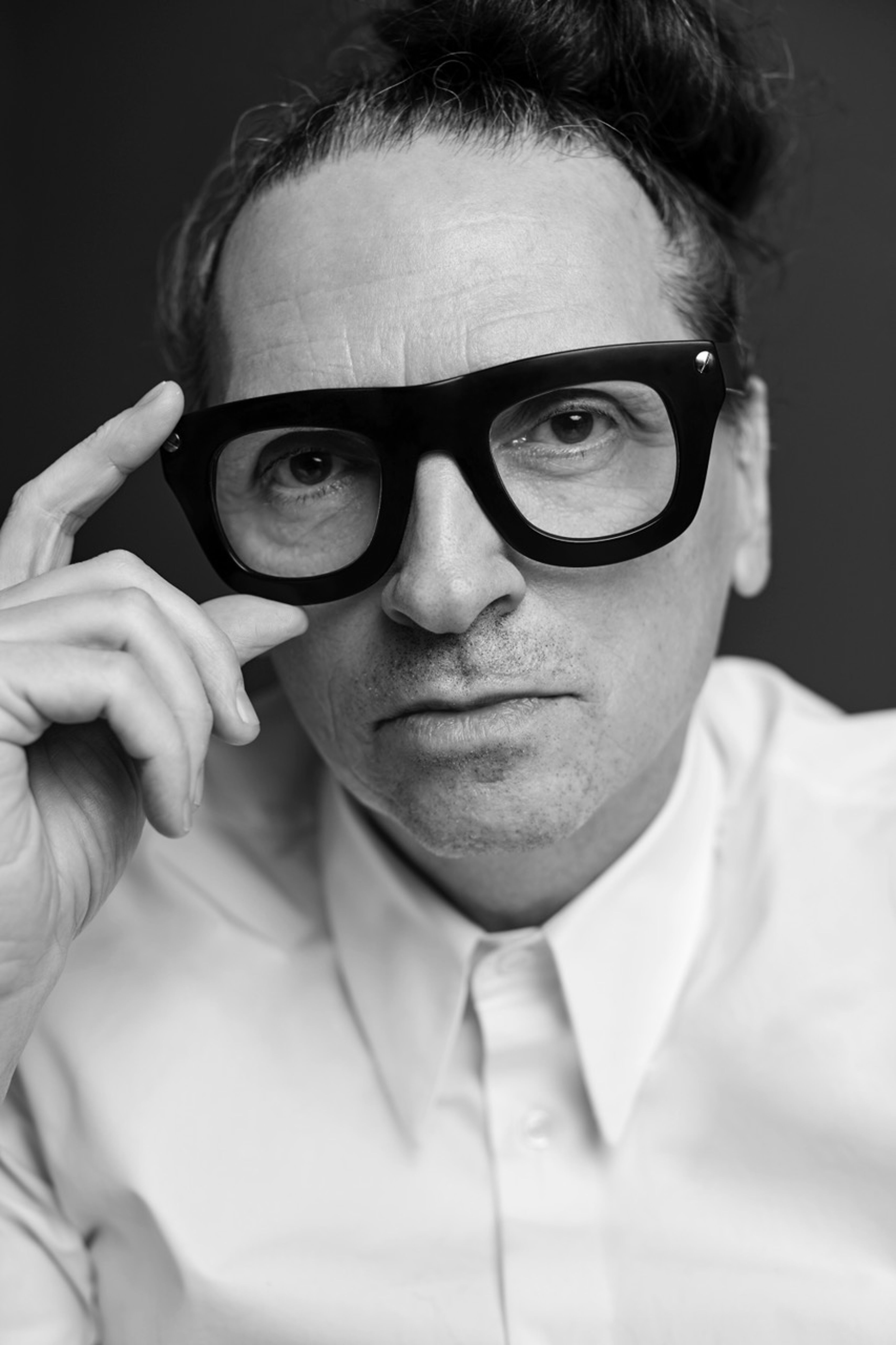 A black and white image capturing a man with distinctive large-framed glasses, looking intently towards the viewer.