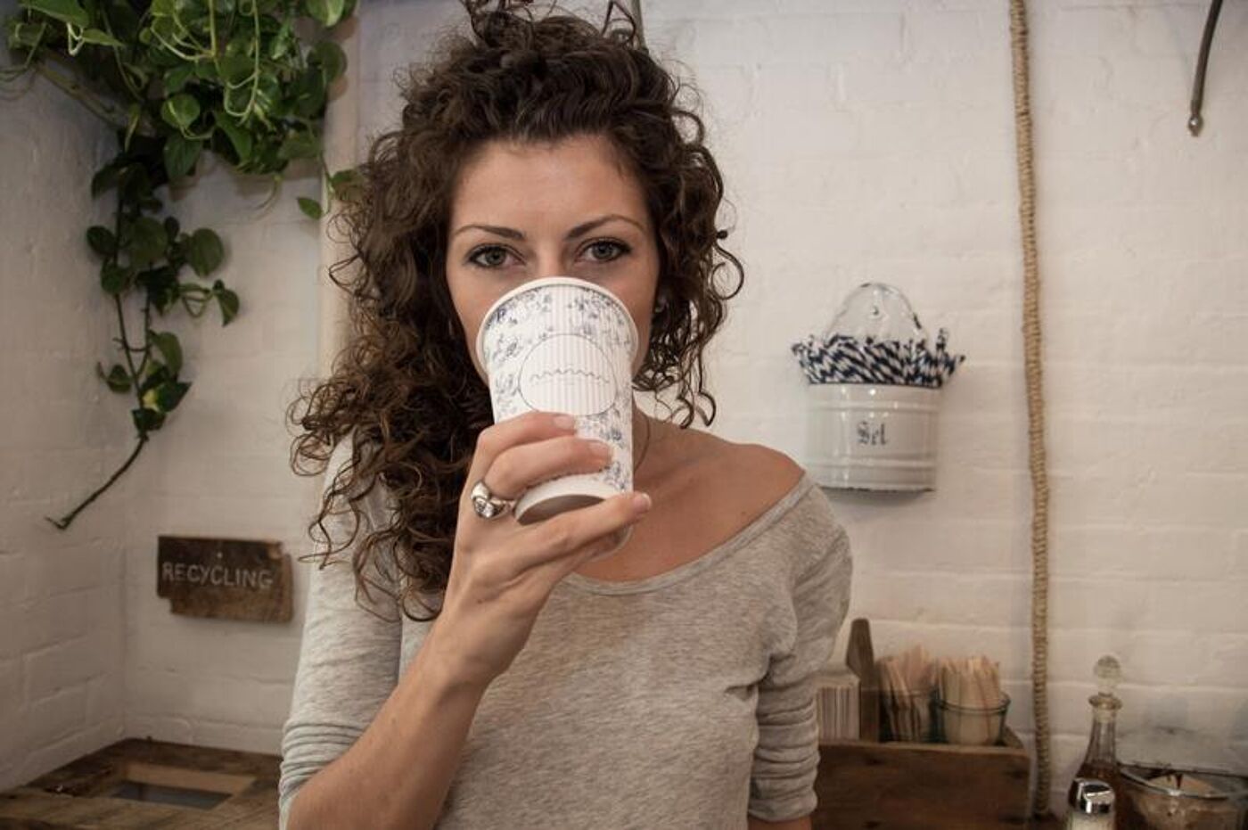 A woman with curly hair drinks from a patterned cup in a rustic kitchen with a recycling sign visible.