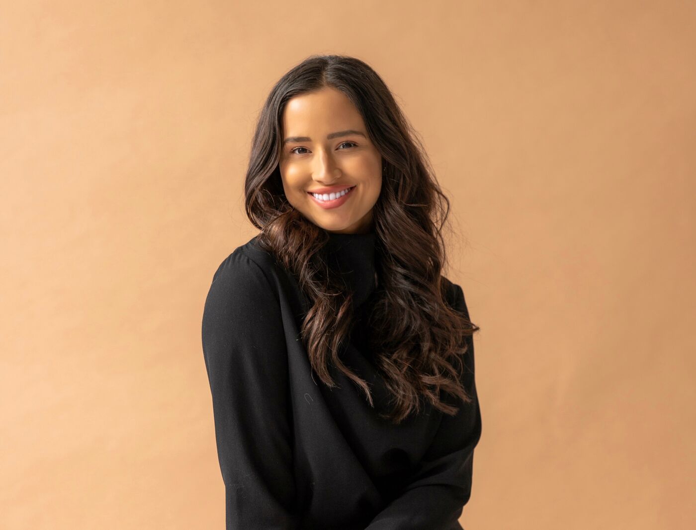 A smiling woman with long, wavy hair sits casually on a stool, wearing a black turtleneck and gray trousers against a warm beige background.
