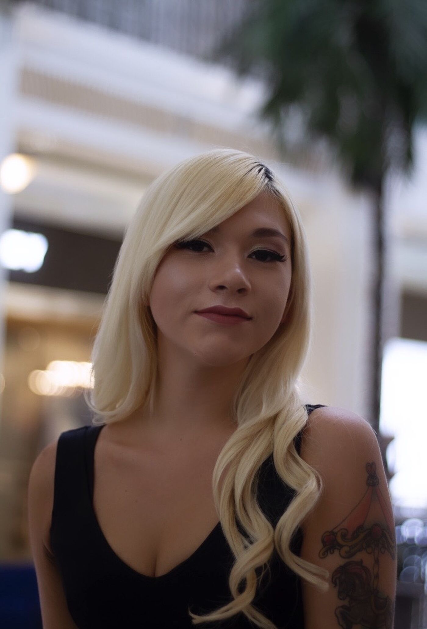 A woman with platinum blonde hair and a visible arm tattoo poses indoors, hinting at a confident personality.