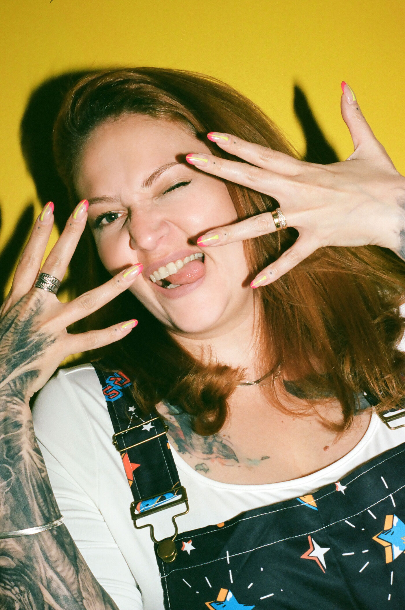 A woman with tattooed arms and vibrant nail art playfully sticks out her tongue, her joyous expression against a yellow backdrop.