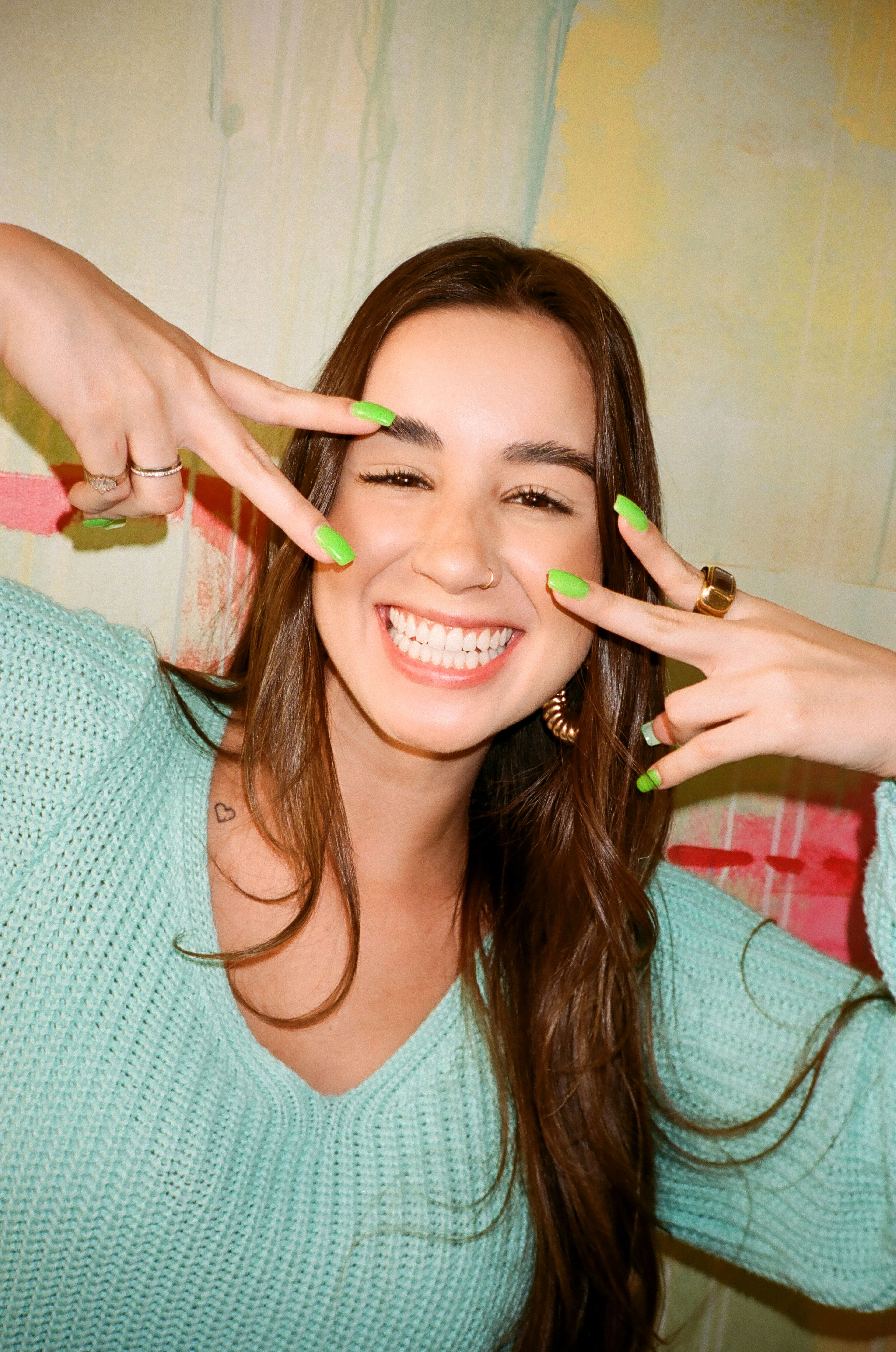 A smiling woman with long brown hair poses playfully, flashing the peace sign near her face, with bright green nail polish, against a softly blurred artistic background.