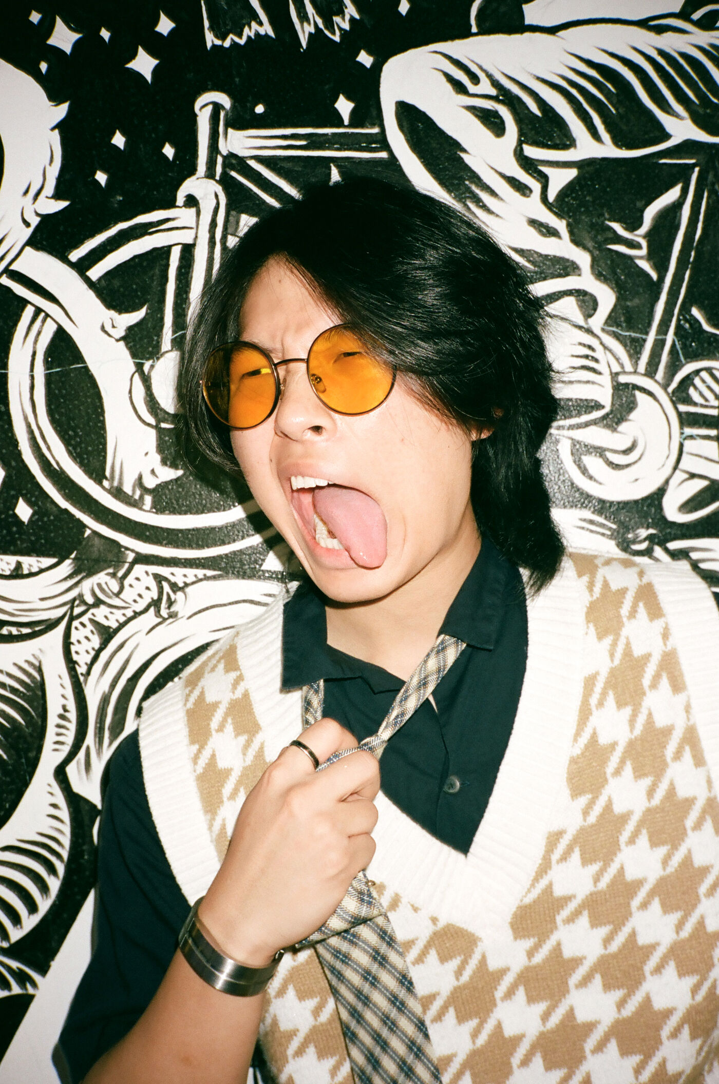 A person in round orange-tinted glasses playfully sticks out their tongue against a black-and-white graphic mural.