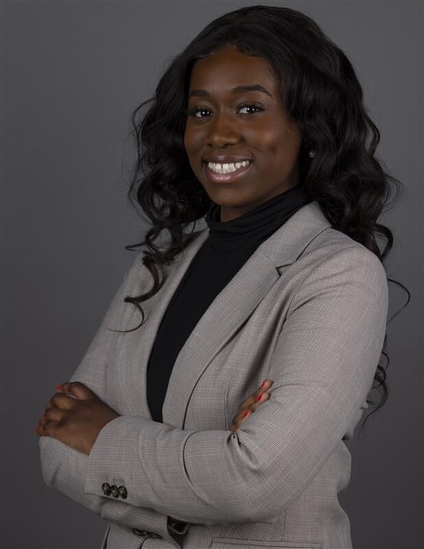A smiling woman with her arms crossed, dressed in a gray blazer and a black turtleneck. She has long, wavy black hair and is looking at the camera.
