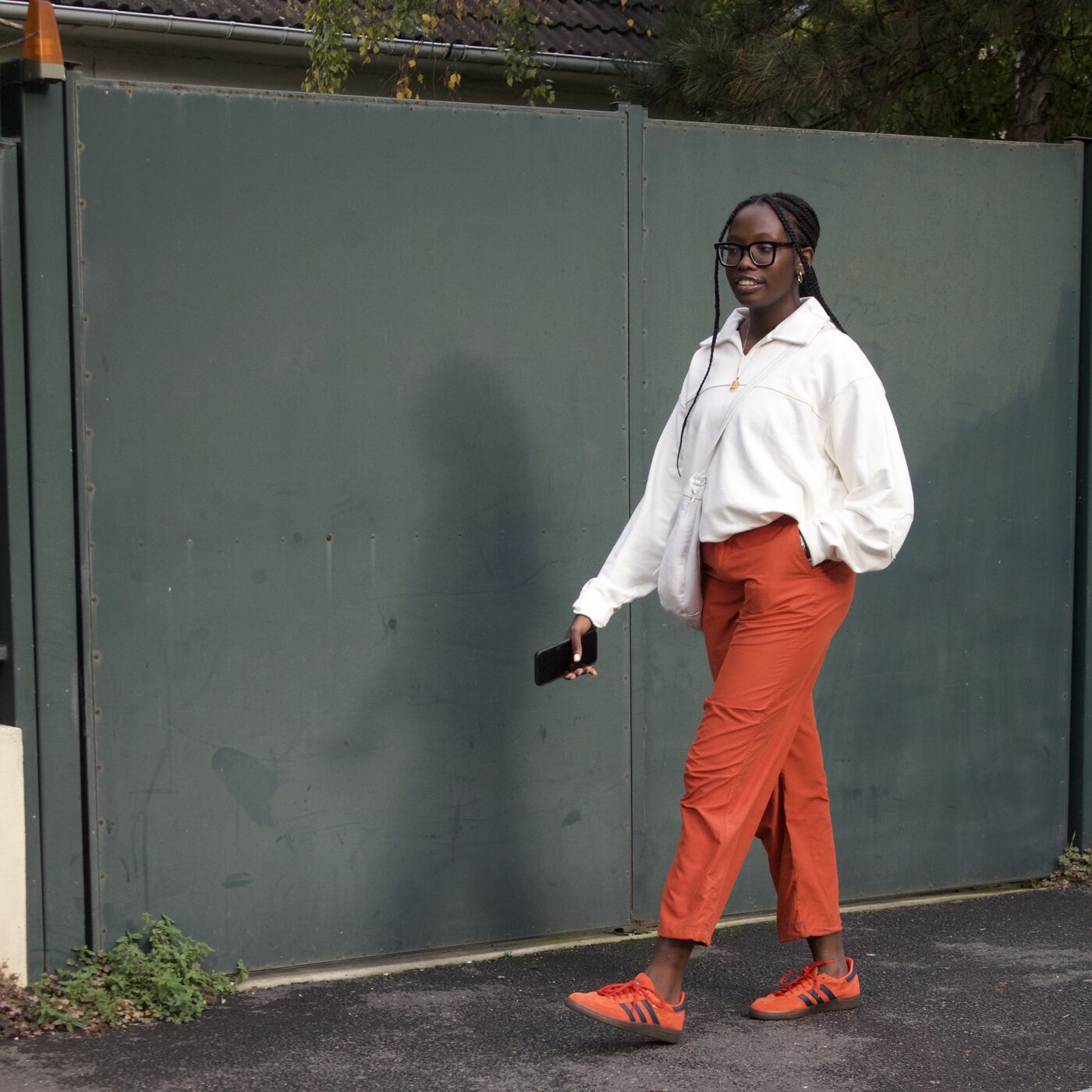 A woman with glasses and braided hair strides confidently by a green gate, sporting a white blouse, orange trousers, and matching sneakers.