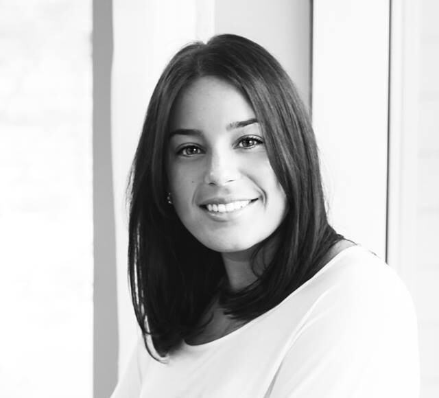 A black-and-white portrait of a smiling woman with crossed arms wearing a white top with button details and a black skirt.