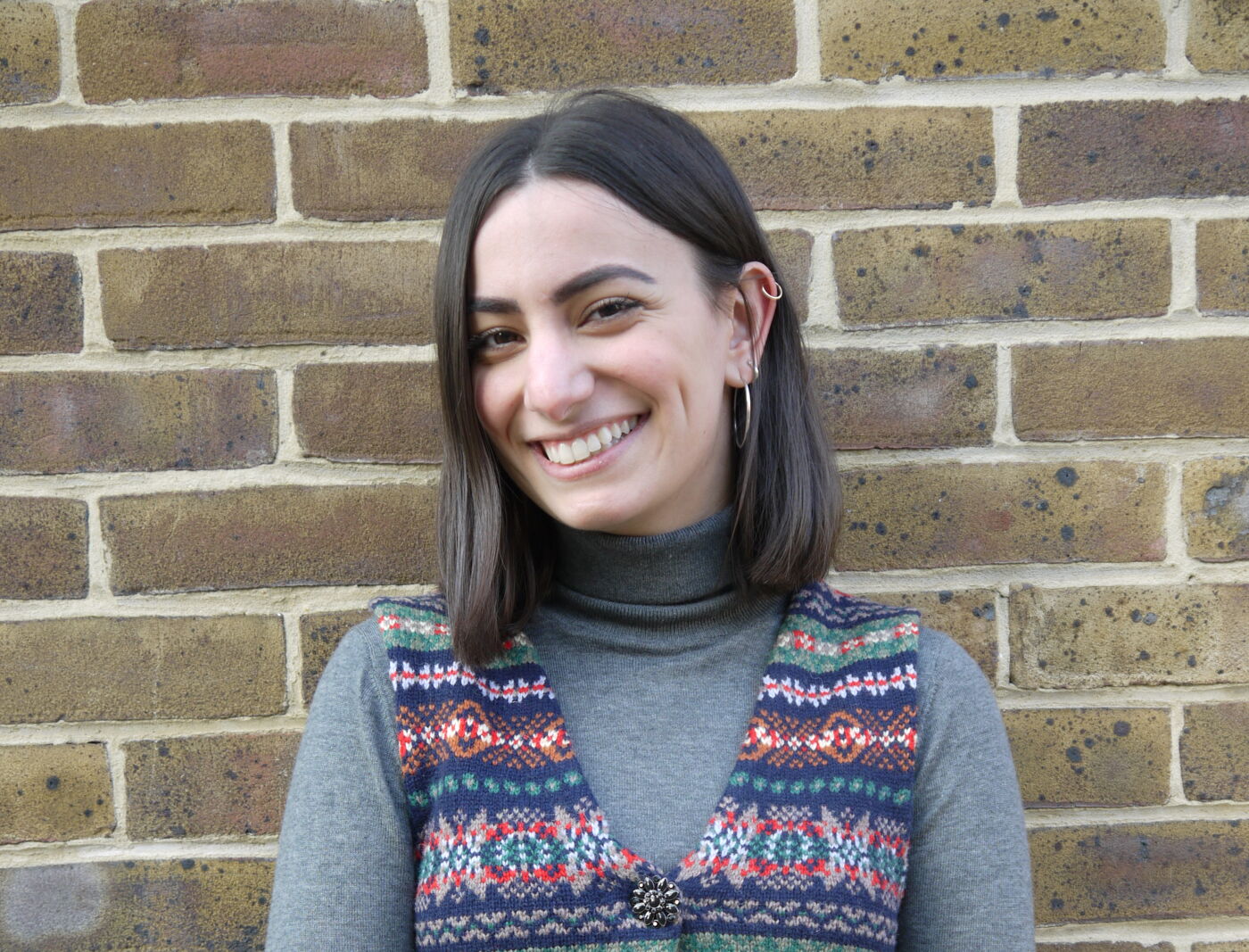 A joyful young woman with shoulder-length hair, wearing a grey turtleneck, a patterned vest, and jeans, standing before a brick wall.