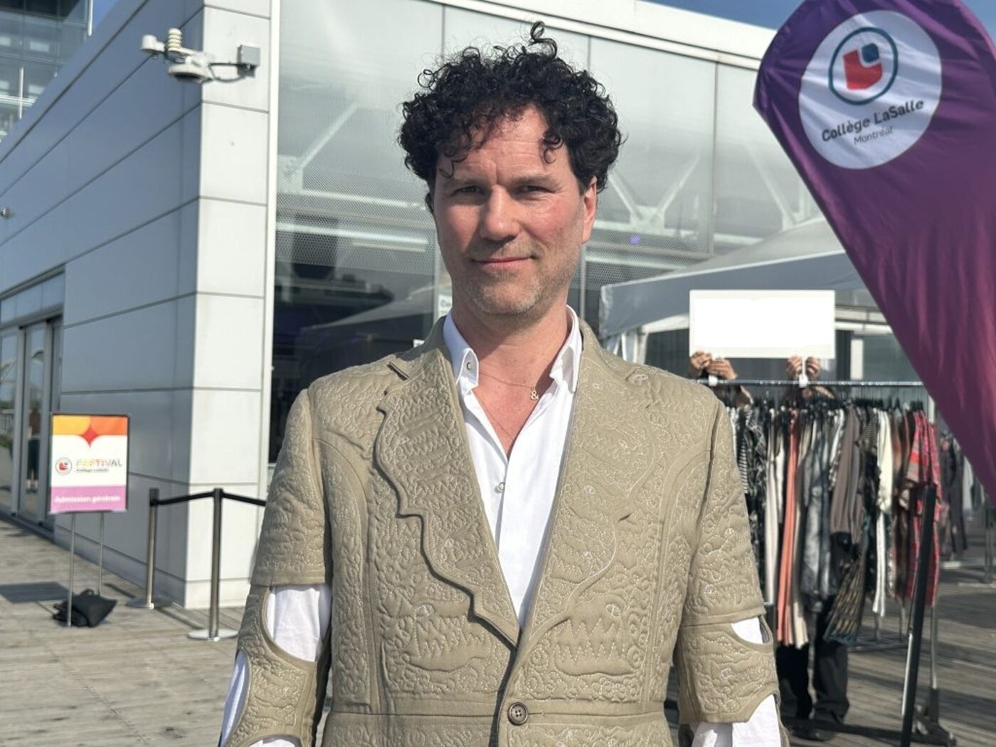 A man with curly hair stands in front of a building, wearing a patterned beige jacket and a white shirt. Behind him, there's a clothing rack with various garments and a purple flag with a logo.