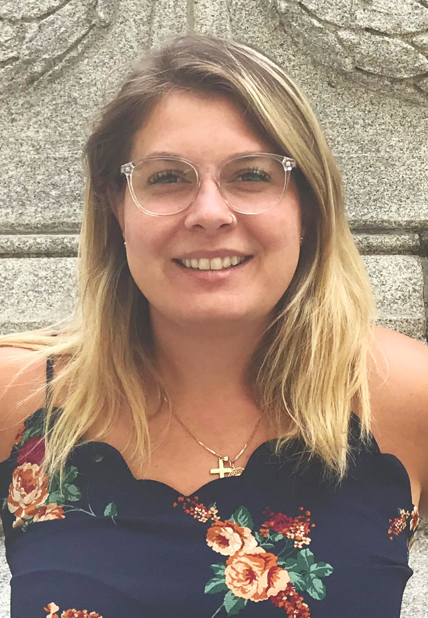 A cheerful woman with shoulder-length blonde hair, wearing clear-framed glasses and a navy floral dress, stands in front of a stone wall.