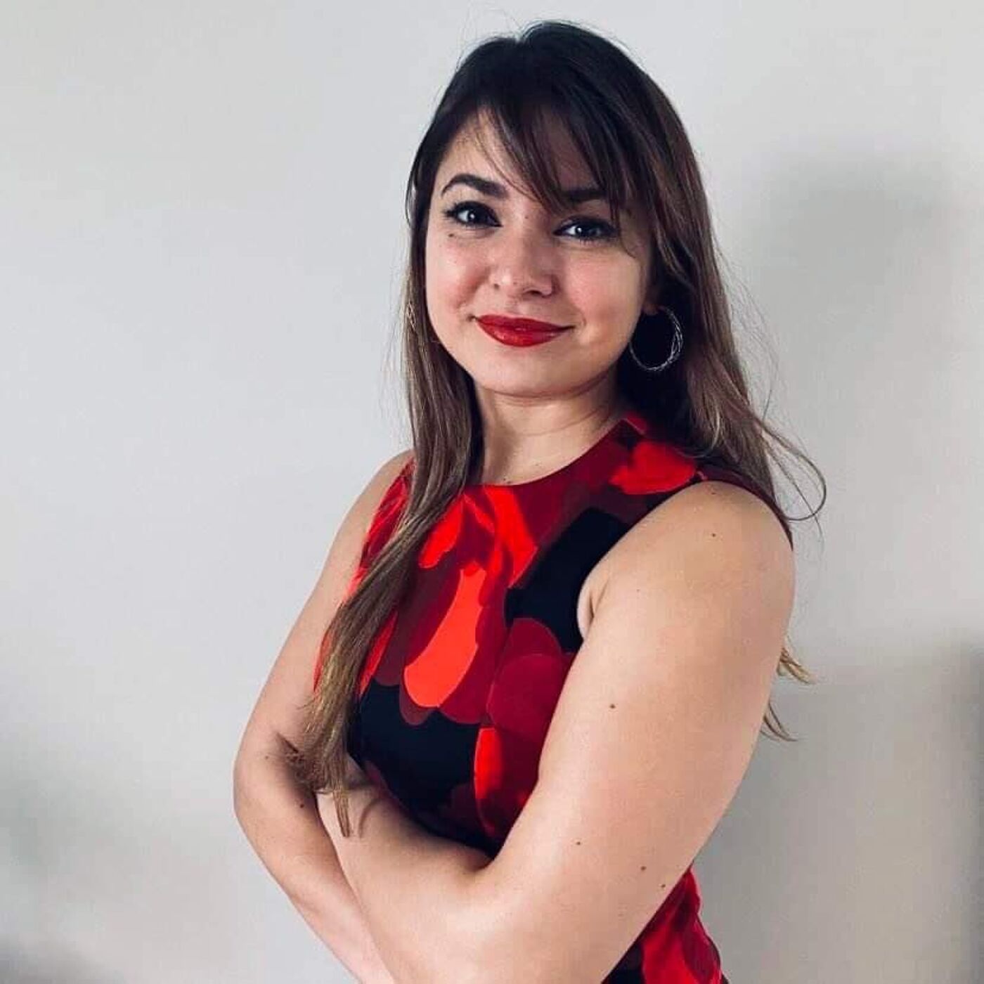 A woman with a poised stance and a pleasant smile, wearing a red and black patterned dress with hoop earrings.